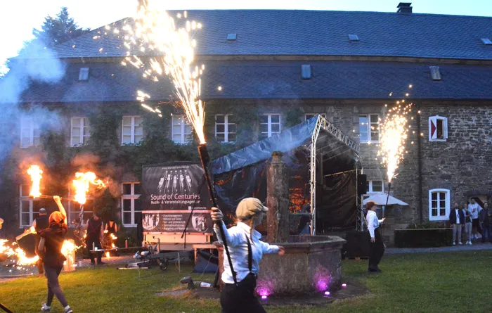 Vor einem historischen Schloss entzünden mehrere Personen mit Fackeln und Feuerwerkskörpern eine Holzkonstruktion, während im Hintergrund weitere Menschen stehen.