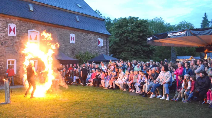 Ein Mann steht inmitten eines großen Feuers vor einer Gruppe von Zuschauern vor einem alten Schlossgebäude.