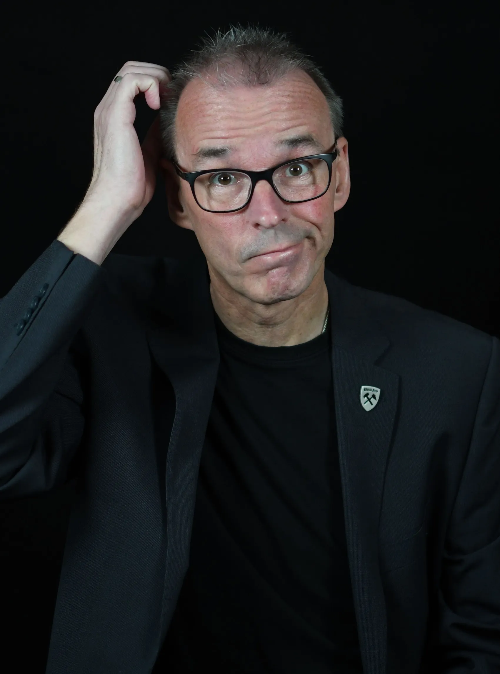 Porträt von Kabarettist Jochen Schmölen mit nachdenklichem Gesichtsausdruck, Hand am Kopf, vor dunklem Hintergrund.