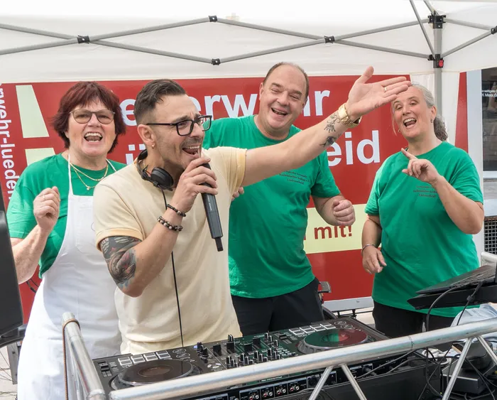 DJ Franco singt in ein Mikrofon und gestikuliert vor einem DJ-Pult, während im Hintergrund drei Personen in grünen T-Shirts mitfeiern.