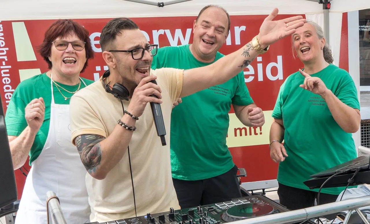 Ein singender Mann mit Mikrofon und ausgestrecktem Arm wird von drei lachenden Personen in grünen T-Shirts begleitet.