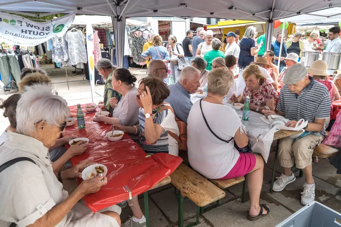 Menschen sitzen unter einem weißen Zelt an langen Tischen mit roten Decken und essen gemeinsam aus Schalen.