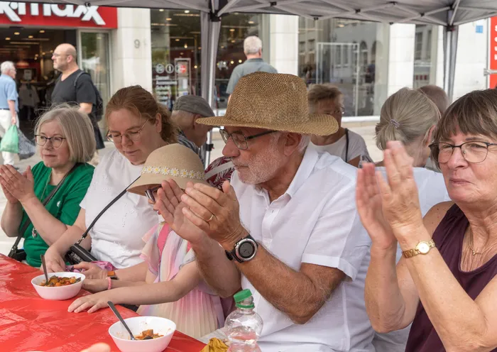 Menschen sitzen an einem Tisch mit roter Decke und klatschen zur Musik, während Schalen mit Caponata vor ihnen stehen.