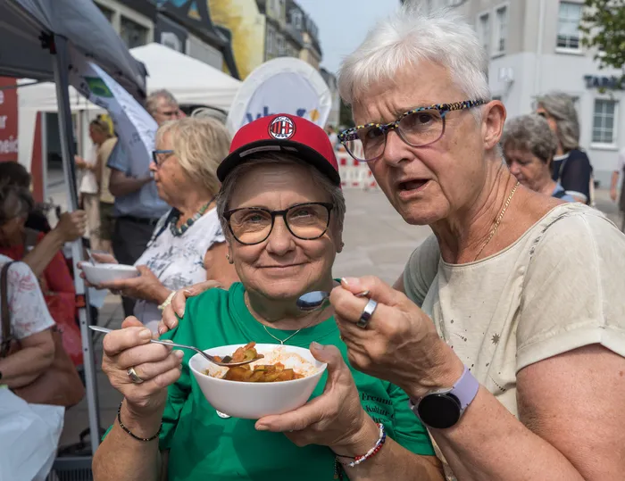 Eine Frau in grüner Kleidung und roter Kappe hält eine Schale mit Suppe, während eine weitere Frau mit Brille daneben steht.