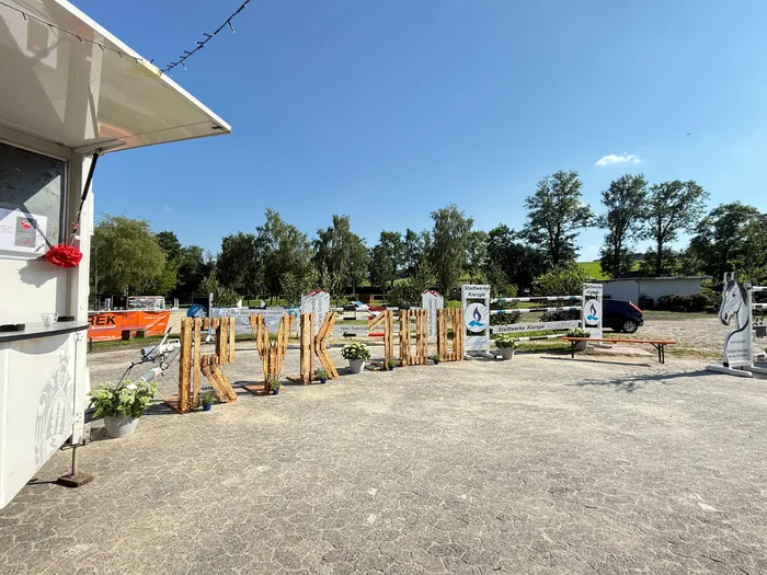 Ein Verkaufsstand steht links im Bild, davor ein grauer Kiesplatz. Im Hintergrund sind Hindernisse für das Reiten aufgebaut, mit Werbebanden für Stadtwerke Kierspe. Bäume und ein blauer Himmel bilden die Kulisse.