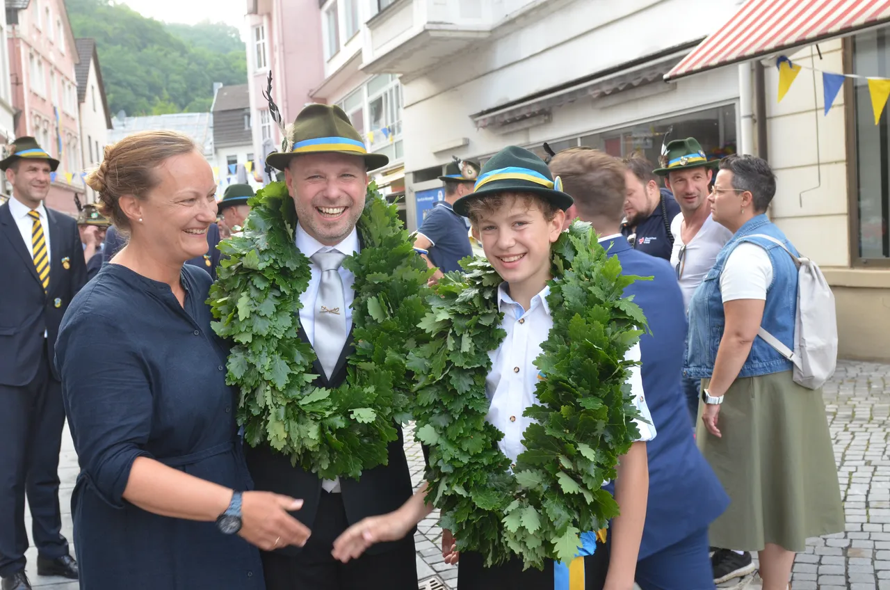 Drei Personen mit großen grünen Eichenlaubkränzen stehen im Mittelpunkt des Bildes. Links eine Frau, in der Mitte ein Mann und rechts ein Junge, alle lächeln in die Kamera. Im Hintergrund sind weitere Menschen und traditionelle Fachwerkhäuser zu sehen.