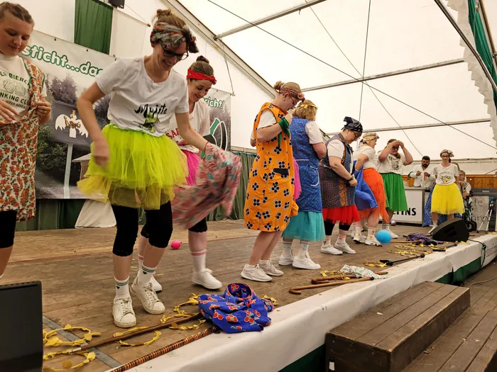 Frauen in bunten Kostümen, Tutus und gemusterten Schürzen stehen auf einer Holzbühne in einem Festzelt.
