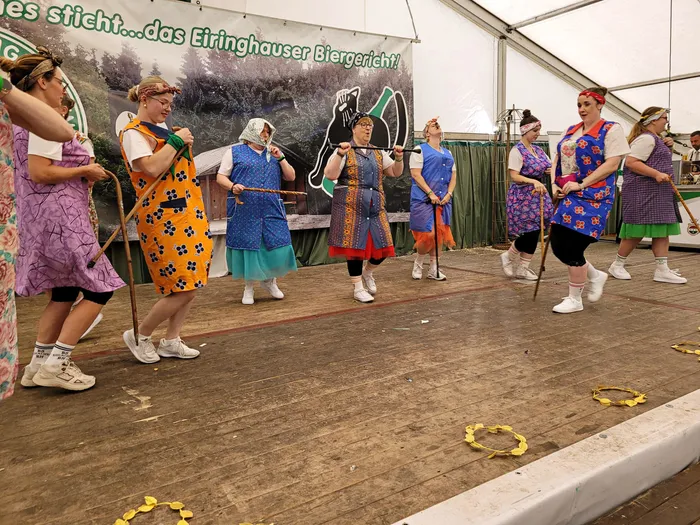 Mitglieder der Biergerichtskommission in bunten, gemusterten Schürzen führen eine Aufführung auf einer Holzstage im Festzelt auf.
