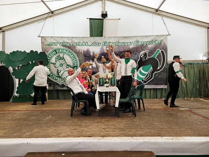 Mitglieder der Biergerichtskommission stoßen auf einer Bühne in einem Festzelt mit Bierkrügen an, vor einem Banner mit der Aufschrift „...Nur eines sticht...das Eiringhauser Biergericht!“.