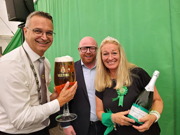 Drei lächelnde Personen in einem Festzelt: Ein Mann hält ein großes Glas Veltins-Bier, eine Frau mit Tiara und grüner Rosette eine Flasche „Schützenkönigin“.