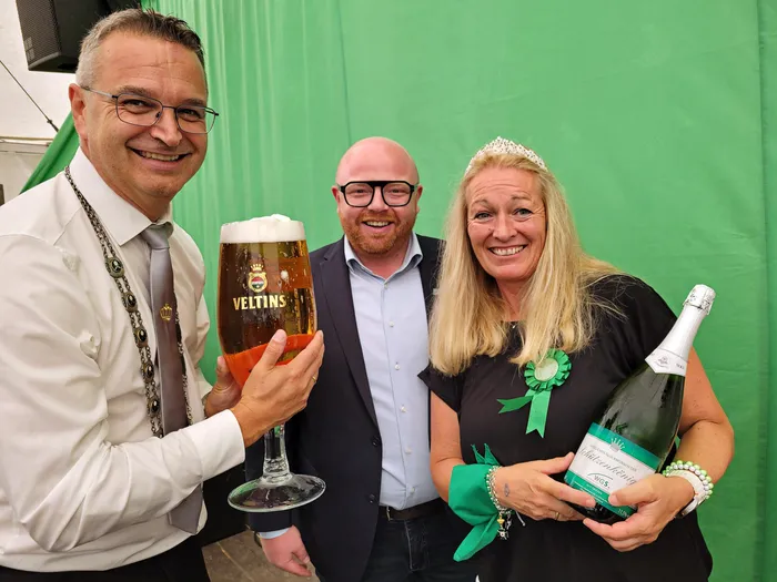 Ein Mann mit Schützenkette hält ein großes Glas Bier, eine Frau mit Tiara und Rosette hält eine Sektflasche vor einem grünen Hintergrund.