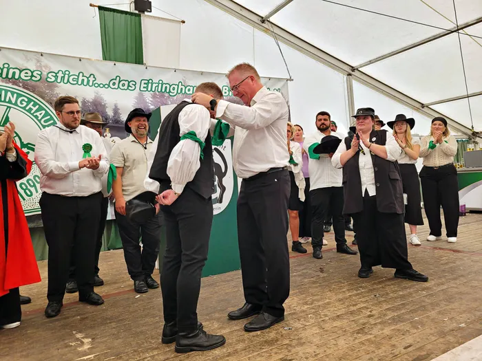 Ein Mitglied der Biergerichtskommission in weißem Hemd und schwarzer Weste wird im Festzelt von einem anderen Mann bei seiner Kleidung unterstützt.