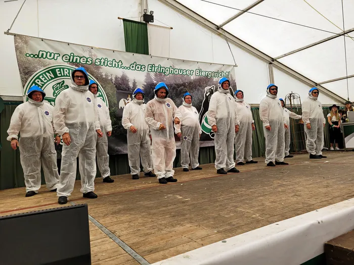 Mitglieder der Biergerichtskommission in weißen Schutzanzügen stehen auf einer Bühne vor einem Banner mit der Aufschrift „...nur eines sticht...das Eiringhauser Biergericht!“.