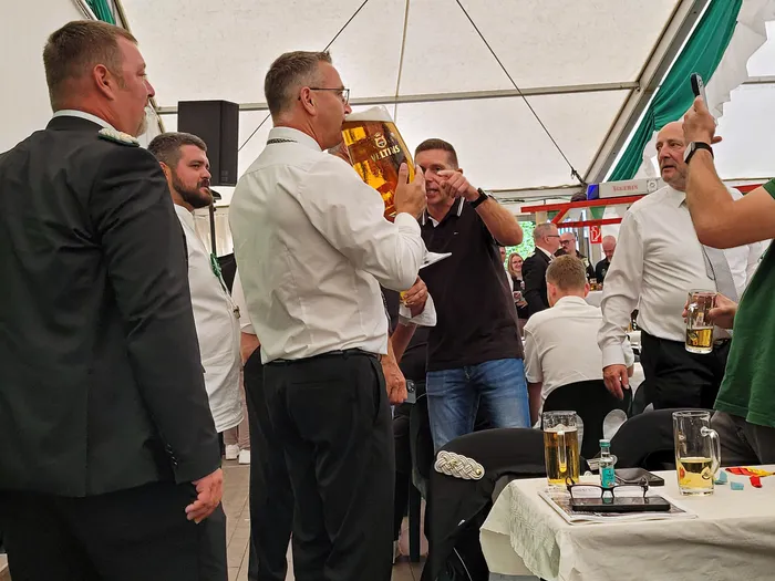 Ein Mann in einem weißen Hemd hebt einen großen Bierkrug hoch, während ein anderer Mann daneben zeigt, im Rahmen der Biergerichtskommission.