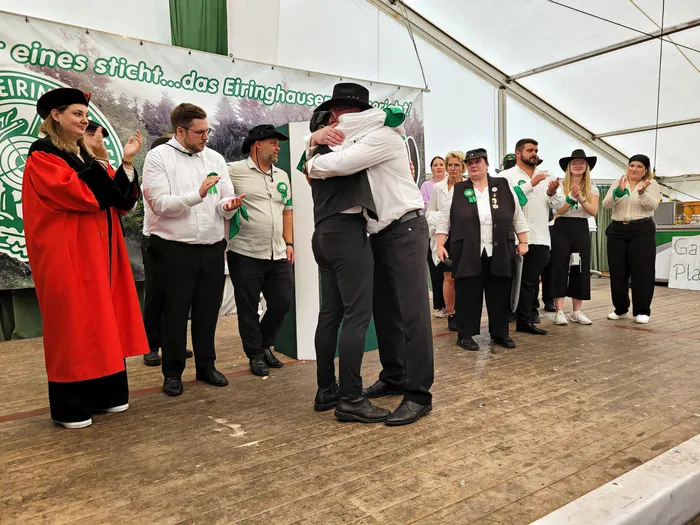 Zwei Männer umarmen sich auf einer Bühne im Festzelt vor einer Gruppe von Menschen in festlicher Kleidung.