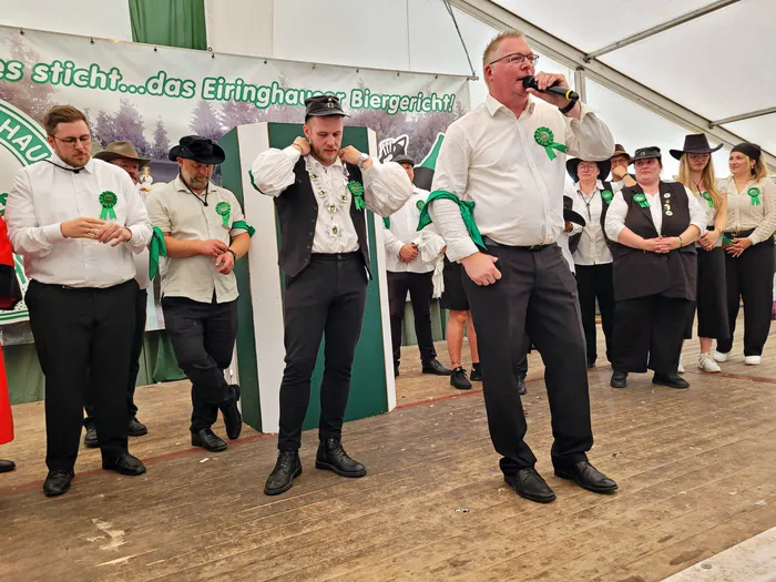 Ein Mann spricht in ein Mikrofon auf einer Bühne im Festzelt, umgeben von Mitgliedern der Biergerichtskommission mit grünen Rosetten.