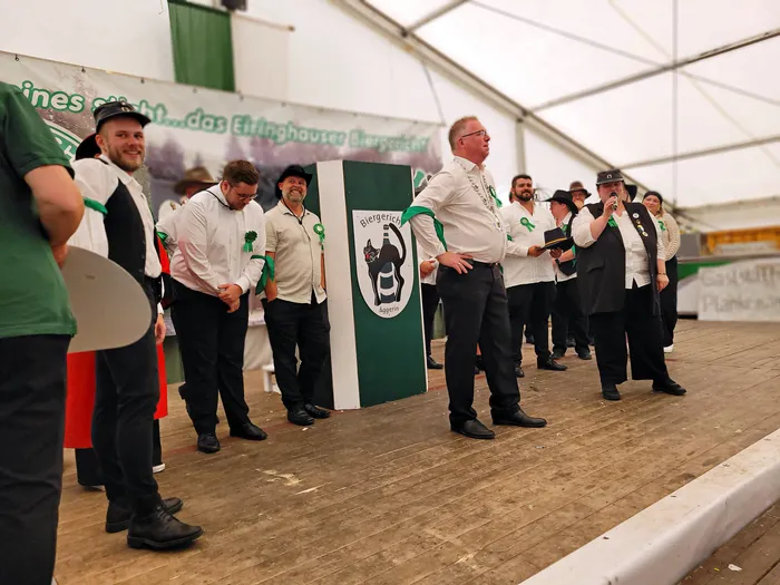 Mitglieder der Biergerichtskommission in festlicher Kleidung stehen auf einer Bühne in einem Festzelt, während ein Mann in ein Mikrofon spricht.