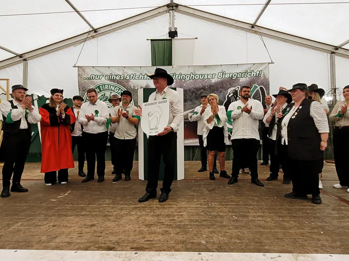 Mitglieder der Biergerichtskommission stehen auf einer Bühne im Festzelt, während ein Mann in der Mitte ein Schild mit der Aufschrift 'Biergericht Eiringhausen' hochhält.