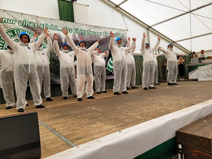 Mitglieder der Biergerichtskommission in weißen Schutzanzügen stehen mit erhobenen Armen auf einer Bühne im Festzelt.
