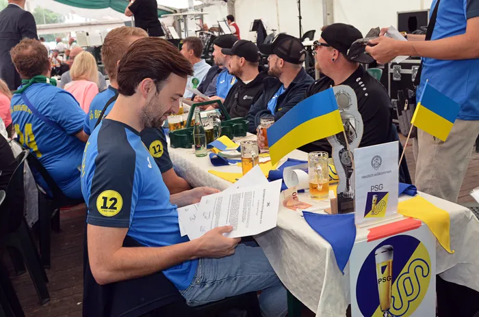 Männer in blauen T-Shirts sitzen an einem Tisch in einem Festzelt mit Biergläsern, Papieren und kleinen ukrainischen Flaggen.