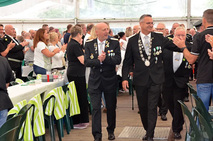 Männer in Schützenuniformen mit Medaillen und silbernen Ketten treten in einem Festzelt vor Zuschauern auf.