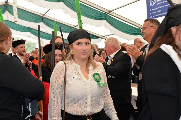 Eine Frau in weißer Spitzenbluse und schwarzem Kopftuch steht im Vordergrund eines Festzeltes, umgeben von Menschen in festlicher Kleidung und Uniformen.