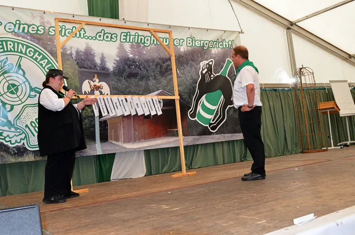 Zwei Personen der Biergerichtskommission stehen auf einer Bühne in einem Festzelt vor einem Banner und einem Holzrahmen.