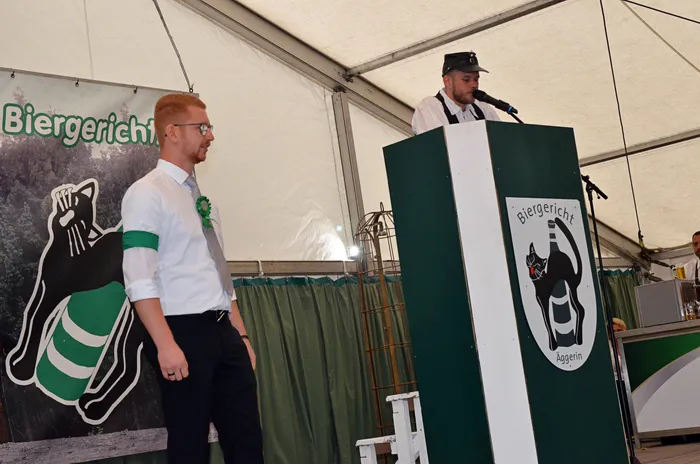 Ein Mann in weißem Hemd steht neben einem grünen Rednerpult mit dem Logo „Biergericht Aggerin“, hinter dem ein Sprecher am Mikrofon steht.
