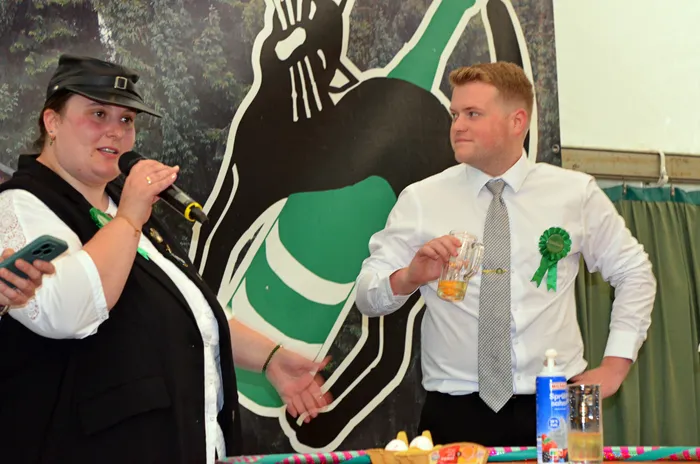 Eine Frau spricht in ein Mikrofon, während ein Mann mit einem Bierglas und einer grünen Rosette neben ihr steht.