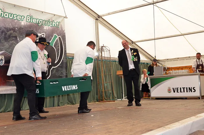 Drei Männer in weißen Hemden mit grünen Ärmeln tragen eine grüne Veltins-Kiste über eine Bühne im Festzelt, während ein Mann im Anzug zusieht.