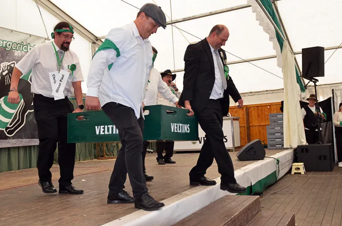 Mitglieder der Biergerichtskommission tragen grüne Veltins-Bierkisten über eine Bühne im Festzelt.