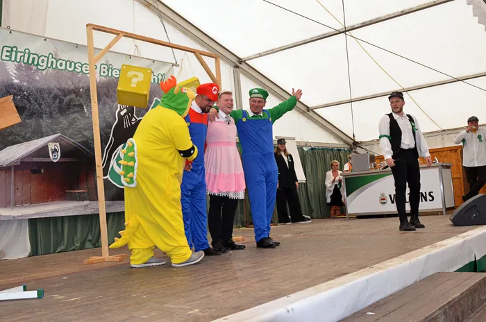 Personen in bunten Kostümen, darunter ein gelbes Dinosaurier-Kostüm, stehen auf einer Bühne in einem Festzelt.