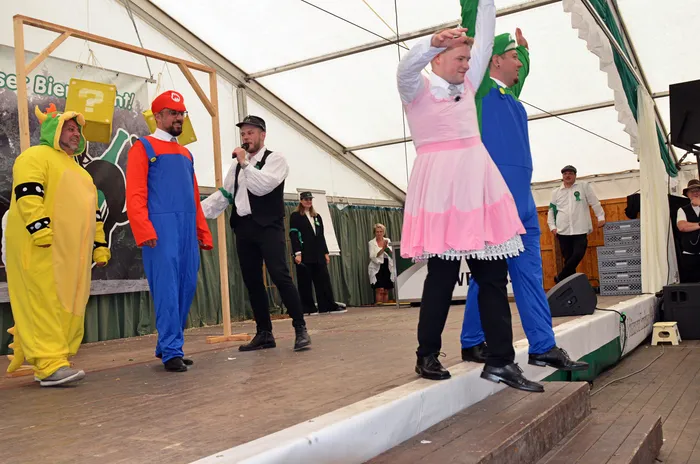 Personen in bunten Kostümen, darunter ein gelber Dinosaurier-Anzug und ein Mann in einem rosa Kleid, treten auf einer Bühne in einem Festzelt auf.