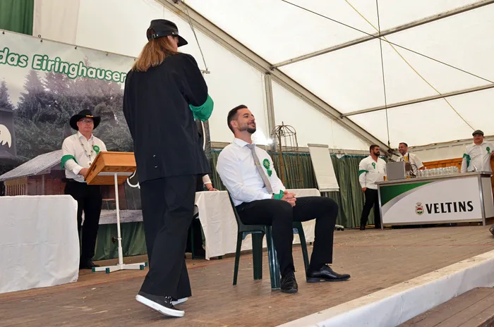 Eine Person in schwarzer Kleidung steht einem sitzenden Mann in weißem Hemd in einem Festzelt gegenüber. Im Hintergrund stehen Personen an einem Tisch mit der Aufschrift 'VELTINS'.