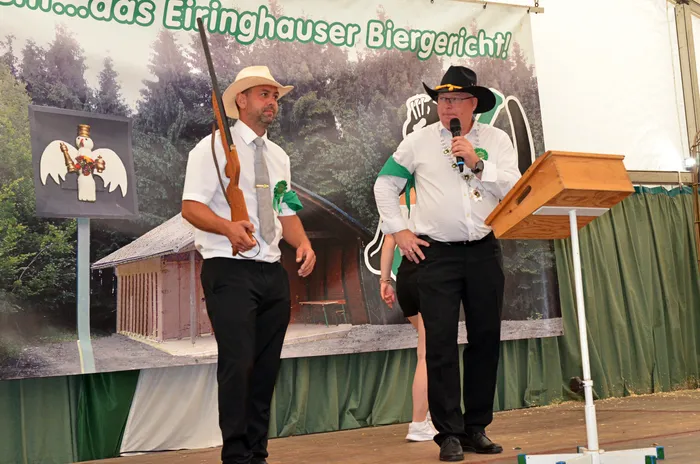 Zwei Männer auf einer Bühne vor einem Banner: Einer mit Strohhut hält ein Gewehr, der andere mit schwarzem Cowboyhut spricht in ein Mikrofon.
