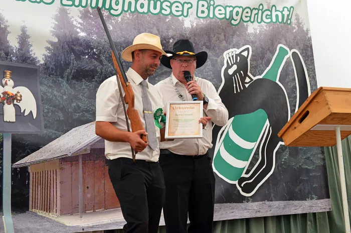 Zwei Männer der Biergerichtskommission stehen vor einem Banner; einer hält ein Mikrofon und eine Urkunde, der andere eine Schrotflinte.