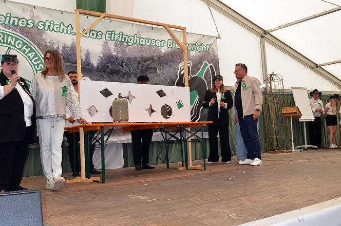 Mitglieder der Biergerichtskommission stehen auf einer Bühne vor einem Holzgestell mit einer weißen Tafel mit rautenförmigen Ausschnitten.