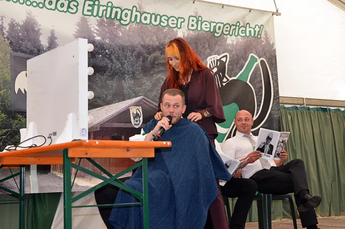 Eine Frau mit roten Haaren steht hinter einem in eine blaue Decke gehüllten Mann mit Mikrofon. Daneben sitzt ein Mann und liest eine Zeitung.