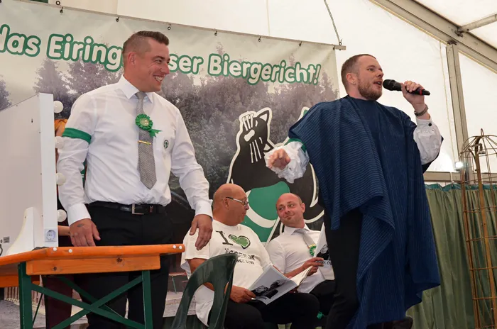 Ein Mann in einem blauen Umhang spricht in ein Mikrofon, während ein Mann in weißem Hemd und mit grüner Rosette lächelnd daneben steht.