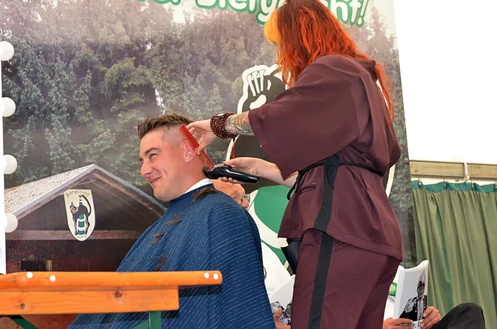 Eine Person mit orangefarbenen Haaren schneidet einem lächelnden Mann im blauen Umhang die Haare.