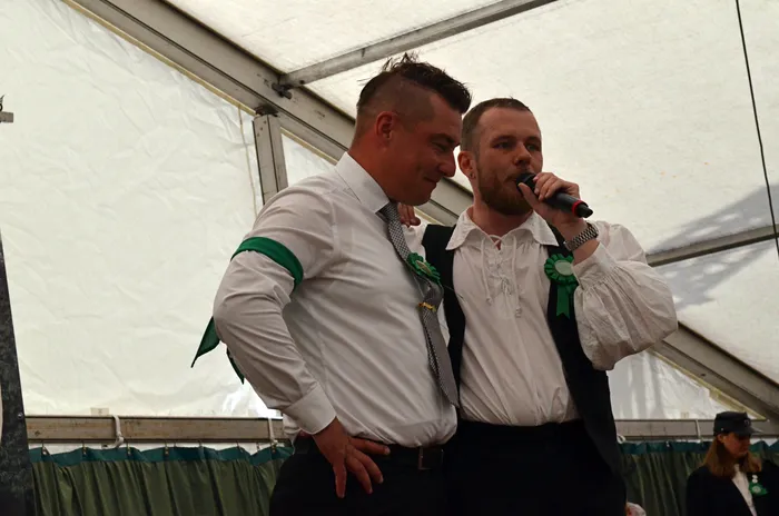 Zwei Männer mit grünen Rosetten stehen in einem Festzelt, einer spricht in ein Mikrofon.