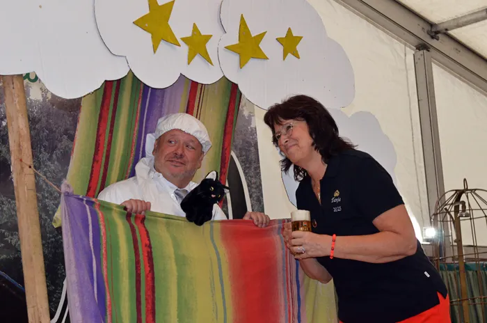 Ein Mann in weißer Kochkleidung und eine Frau mit einem Bierglas stehen vor einer Kulisse mit Wolken und Sternen in einem Festzelt.