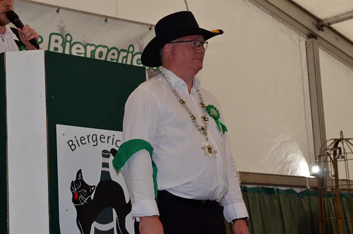 Ein Mann mit schwarzem Hut und einer zeremoniellen Kette steht neben einem Podium mit dem „Biergericht“-Logo.