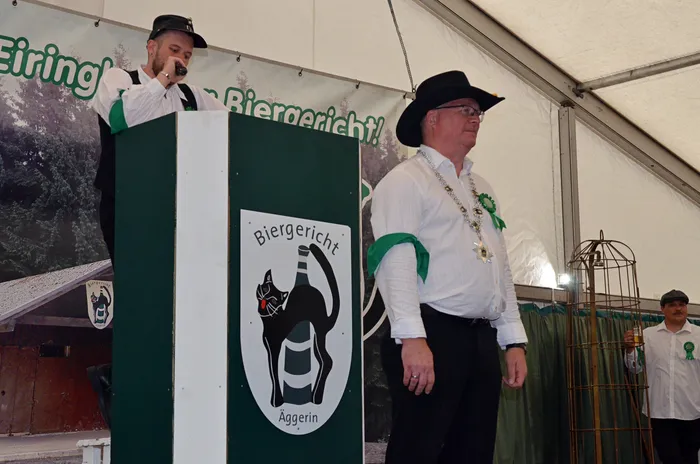 Ein Mann spricht an einem Pult mit dem Logo des „Biergericht Äggerin“, daneben steht ein Mann in weißem Hemd und schwarzem Hut in einem Festzelt.