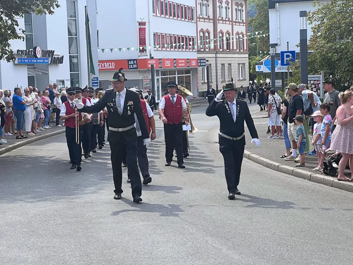 Männer in dunklen Uniformen und Hüten mit Orden marschieren bei einem Festumzug durch eine von Zuschauern gesäumte Straße.
