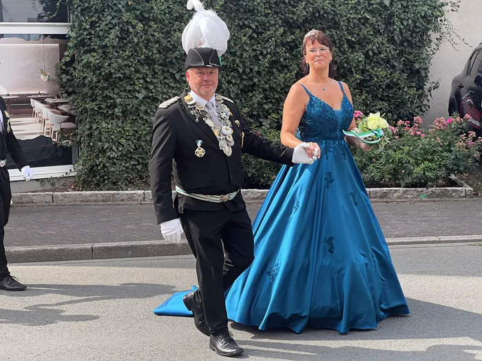 Ein Mann in schwarzer Schützenuniform mit weißem Federhut und eine Frau in einem langen blauen Kleid gehen Hand in Hand bei einem Festumzug.