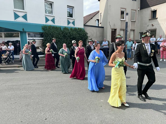 Frauen in farbenfrohen Abendkleidern und Männer in Uniformen ziehen bei einem Schützenfest-Umzug durch die Straße.
