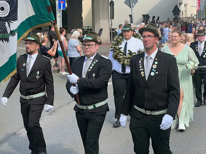 Männer in schwarzen Anzügen, grünen Hüten und weißen Handschuhen ziehen bei einem Festumzug mit einer grün-weißen Fahne durch die Straße.