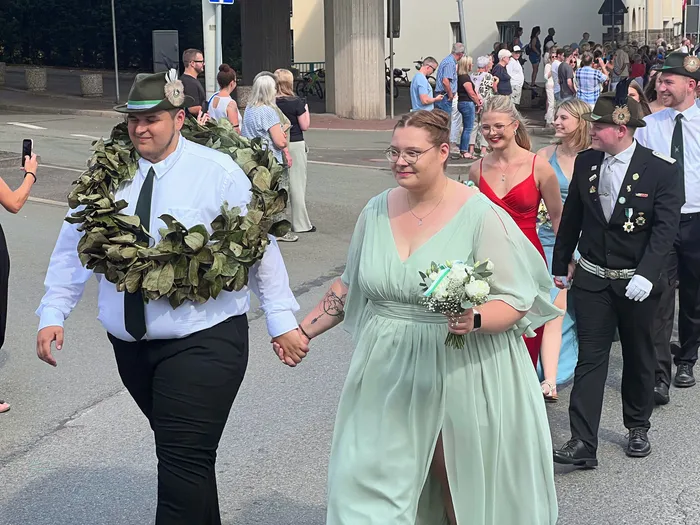 Ein Mann mit einem großen Laubkranz und eine Frau in einem hellgrünen Kleid mit Blumenstrauß gehen Hand in Hand bei einem Festumzug.