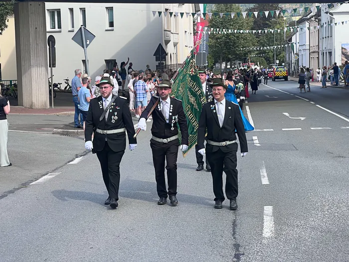 Drei Männer in schwarzen Uniformen mit weißen Gürteln marschieren bei einem Festumzug durch eine Straße, einer trägt eine große grüne Fahne mit goldener Schrift.
